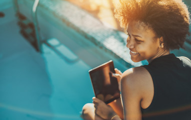 Smiling young beautiful Brazilian female with curly afro hair is sitting on the edge of small swimming pool and holding her digital pad; copy space for text or your advertising; teal and orange colors