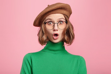 Horizontal shot of astonished beautiful female model wears green turtleneck sweater and glasses, looks with bugged eyes and widely opened mouth, surprised to observe terrified scene on street