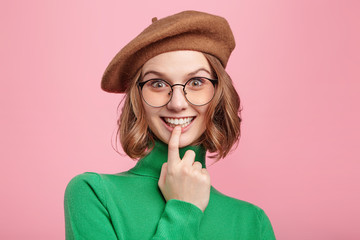 Pleasant looking young female nerd wears spectacles, brown beret and casual green sweater, keeps finger on mouth, glad to recieve pleasant comments of dish she prepared by herself, feels proud - obrazy, fototapety, plakaty