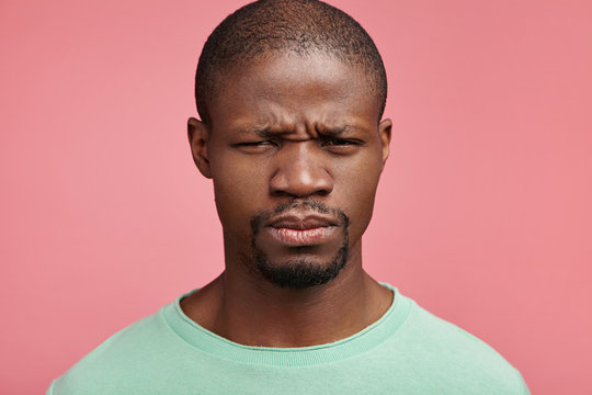 Headshot of sullen moody glum African American man frowns face, being very obused or displeased after conversation with boss. Unhappy upset discontent dark skinned man isolated on pink background