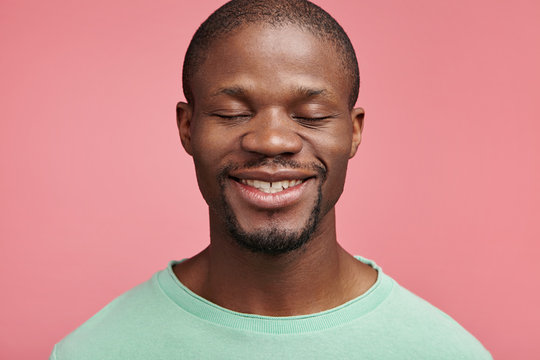 Headshot of glad cheerful dark skinned man closes eyes, smiles happily, going to recieve present, celebrates his work promotion or anniversary, pleased to hear positive words and congratulations