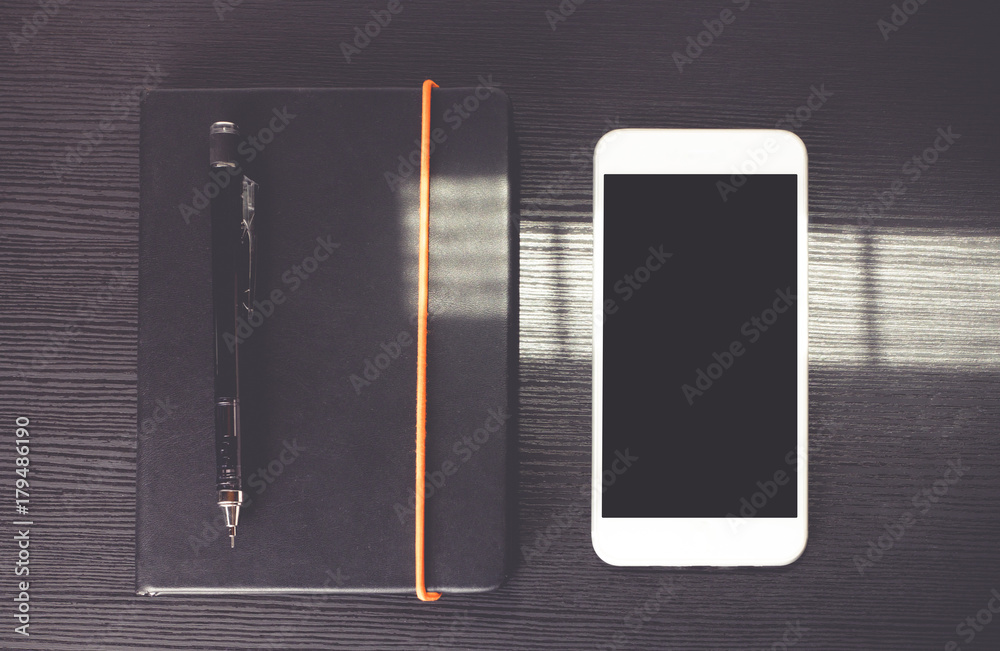 Poster Blank screen of mobile phone lay on black notebook near window on black wooden modern desk at afternoon sunlight, Mock up for display of design or content,Clipping path smart phone screen.