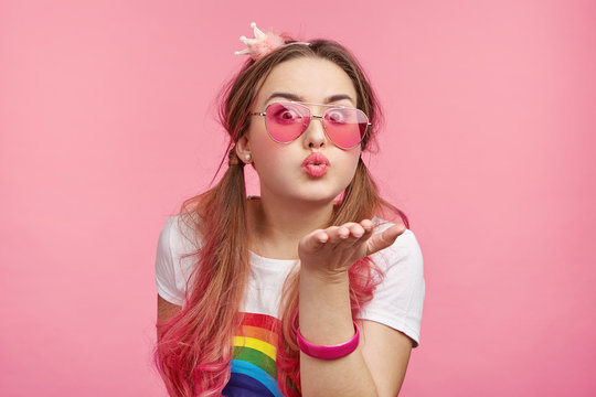 Adorable attractive glamour young woman in round pink glasses sends kiss against pink background. Coquette female blows sweet kiss to boyfriend, flirts. Fashion, feelings and people concept.