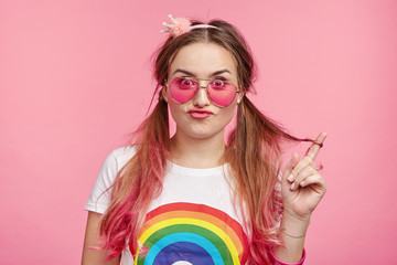 Stylish female model in pink sunglasses and t shirt with rainbow print, makes grimace, curves lips and shows her pony tails, poses against studio background. People, beauty, lifestyle concept