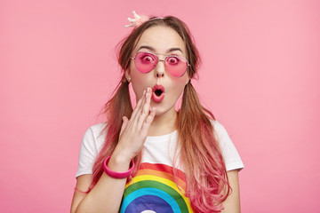 Stylish pin up girl wears pink glasses, has long hair tied in pony tails, expresses disbelief as...