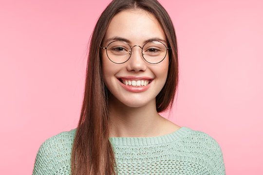Young Asian woman in round spectacles, smiles happily as recieves compliment from stranger, wears casual knitted sweater, isolated over pink background. People, joy, happiness, emotions concept