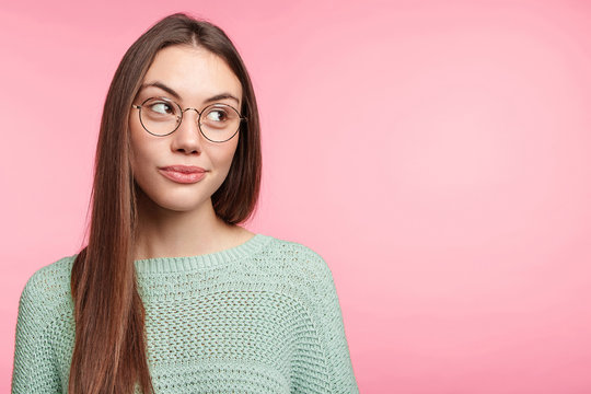 Brunette woman with long hair, wears spectacles and loose sweater, looks thoughtfully aside, generates new ideas for project work, isolated over pink wall with copy space for your advertisment