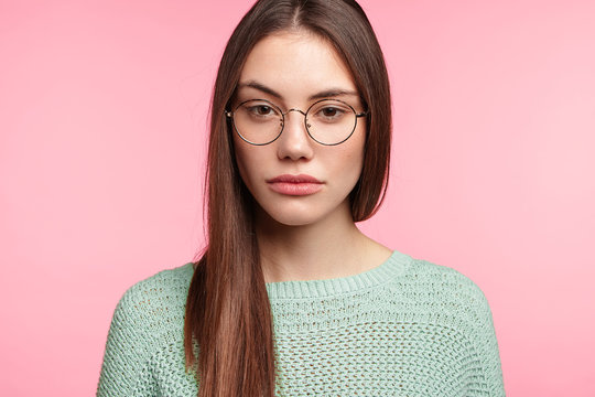 Portrait of attractive young female with Asian appearance wears spectacles, loos seriously at camera, has sad expression, wants sleep, listens to lecture attentively, isolated over pink background