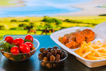 Meat, French fries in a one-time lunch box made of foamed polystyrene, tomatoes with greens and olives on a background of landscape