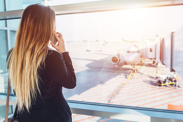 Young businesswoman talking on mobile phone waiting for her flight looking at planes through the window standing in departure zone at airport