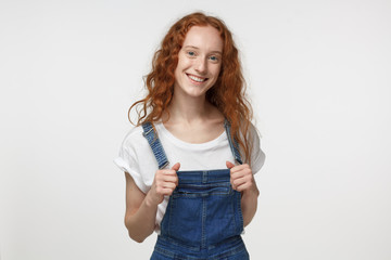 Obraz premium Closeup portrait of young teenage girl with red loose hair pictured isolated on white background pulling forward straps of her denim jumpsuit, smiling happily at camera feeling joy and satisfaction