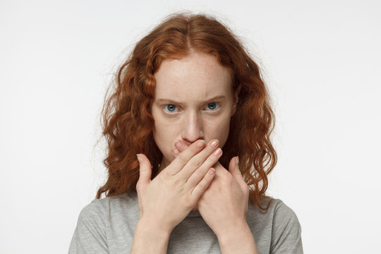Indoor portrait of teenage European girl with red hair pictured isolated on white background, hiding her mouth behind hands in resolute gesture refusing to reveal secrets and share confidential data