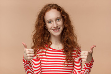 Closeup of young pretty curly redhead girl in striped t-shirt showing thumps up with positive emotions of content and happiness. Concept of satisfaction with quality and recommendation