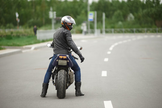 Sports, extreme, speed, adrenaline and determination concept. Rear view of stylish biker in leather clothes and safety helmet riding his motorcycle along empty high way, turning face to camera