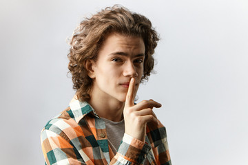 Don't say anybody. Studio shot of cute guy with wavy hair having mournful expression, holding index finger at his lips, asking to keep his secret. Human emotions, feelings, reaction and body language