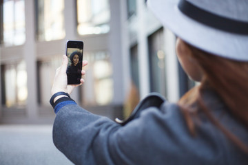Back view. Young modern stylish beautiful woman in hat while walking in the big city streets doing selfie. 