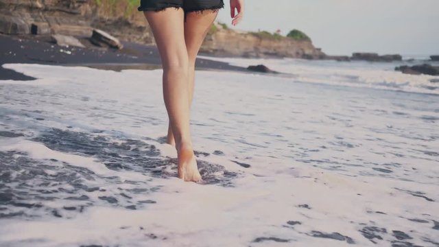Young woman walking along the water's edge. Woman going along the shore on black sand. The camera is moving after the female.