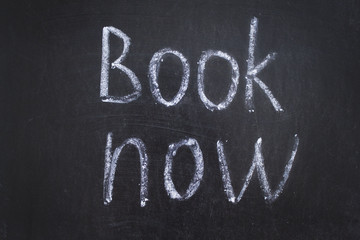 Text Book Now handwritten with white chalk on a blackboard. 