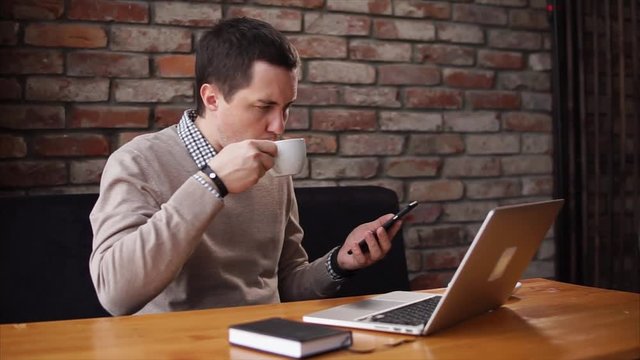 Freelancer is sitting in a caffee with a laptop, smartphone and his diary. He is holdign a smartphone, scrolls the articles and then drink coffee.