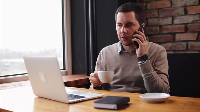 An adult man who drinks hot coffee in a cafe talks on the smartphone with a business partner, on the desk is a laptop for work on the Internet and a notebook for recording notes
