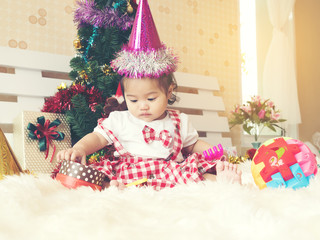 Baby girls on red and white dress and pink hat sitting on white bed with toy and christmas tree on back side