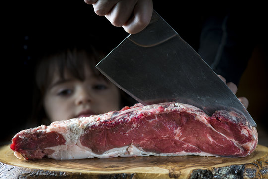Niña pequeña observa con curiosidad a un carnicero cortando entrecot de ternera con una cuchilla.