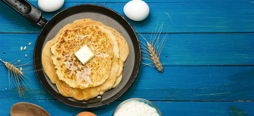 Hot delicious pancakes in frying pan on blue wooden table with flour and eggs. Pancake day background, border design panoramic banner 