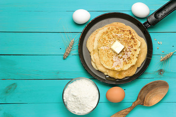 Hot delicious pancakes in frying pan on turquoise wooden table with flour and eggs. Pancake day background