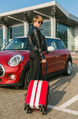 Beautiful girl looking at the camera while staying near the car. Girl is going on vacation with red suitcase. Summer mood, new emotions from future holidays.