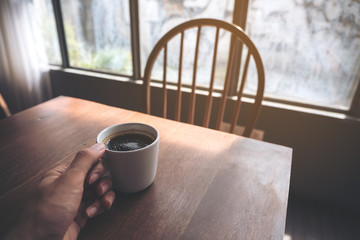 Closeup image of a hand holding a white hot coffee cup on vintage wooden table in cafe