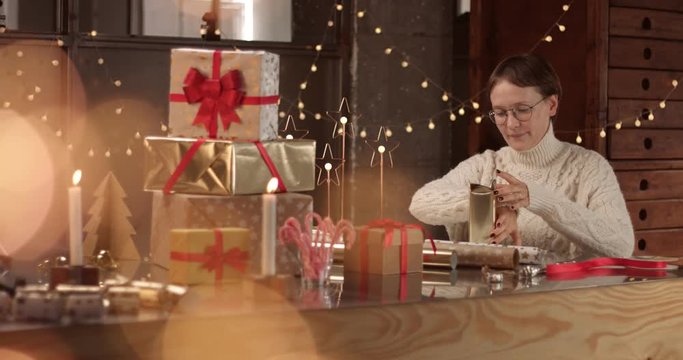 Woman in thick white cable sweater wrapping presents on the background of Christmas decorations and lights, man brings a cup of coffee or hot chocolate