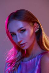 Vertical portrait of young beautiful woman with long hair and professional make up wearing metal accessories and dress, looking in camera with calm expression. Colorful fleshlights. Pink, red, orange