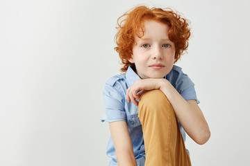 Close up of beautiful ginger boy in blue shirt and yellow jeans holding hand on leg, looking in camera and gently smiling.