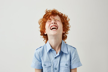 Close up of joyful little kid with ginger curly hair and freckles in blue shirt laughing hard...