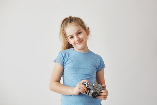 Portrait o good-looking blond child in blue t-shirt smiling, standing with photo camera in hands posing for school album.