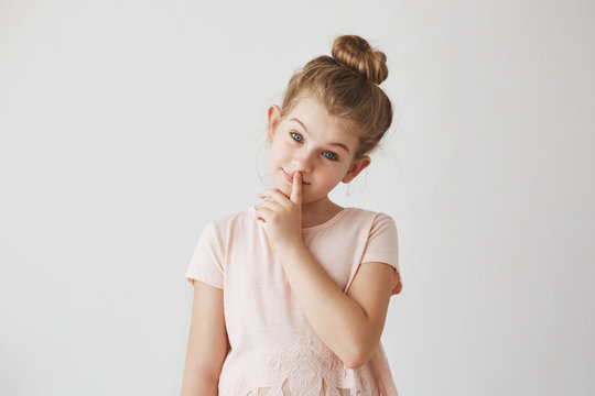 Portrait of tender little girl with blond hair in bun hairstyle , holding finger on lips and smiling, looking in camera with calm face expression.