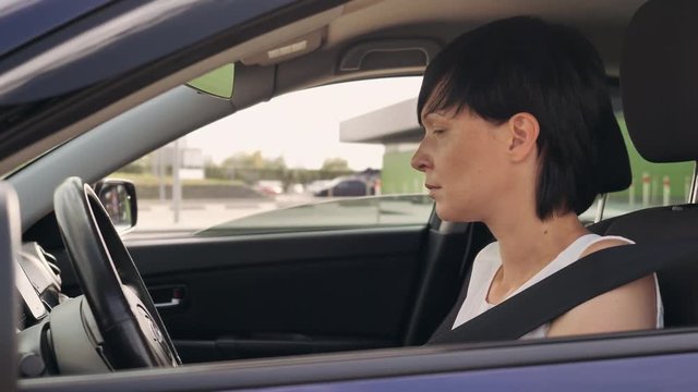 Adult woman sitting in car fastens seat belt. Attractive caucasian model with short black hair leaves the parking lot.