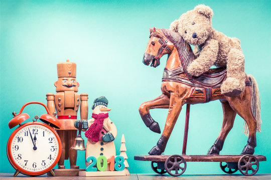 Retro Teddy Bear toy riding antique Christmas wooden horse on wheels, old snowman with jingle bell and 2018 date, nutcracker, little New Year tree, clock. Holiday greeting card. Vintage style photo