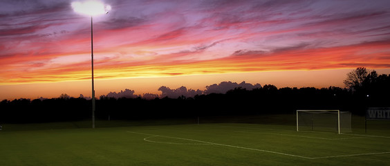 A light towering over an empty soccer field with a sunset in the background - obrazy, fototapety, plakaty