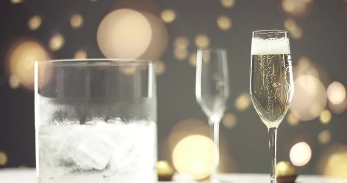 Two glasses of champagne with bubbles and an ice bucket on the background of festive lights and gray wall