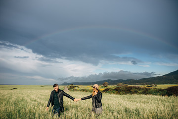 Fototapeta na wymiar Happy loving couple walking around the field holding hands, mountains and rainbow on the background