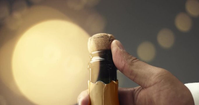Close up slowmotion video of man's hands opening a bottle of champagne on gray background with lights and flares