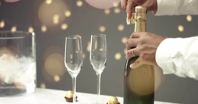 Man in white shirt opens a bottle of champagne at a party on gray background with lights, close up video