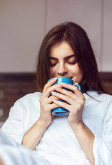 Beautiful caucasian girl, in a white bathrobe, enjoys morning coffee time in the kitchen