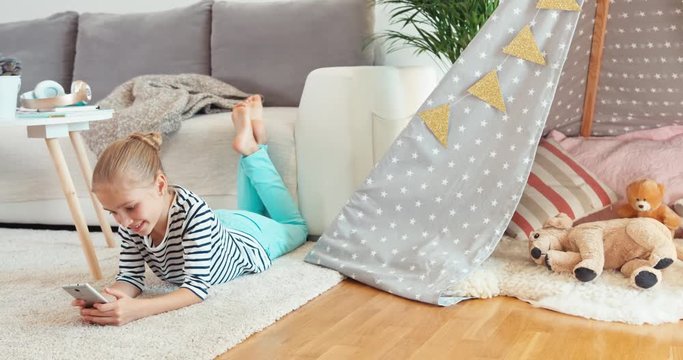 Child girl using cell phone lying on the carpet indoors and smiling at camera