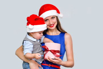 Mother holding her child and shows him a box with a red bow. A little boy takes out the gift from the box. Both in Santa hats