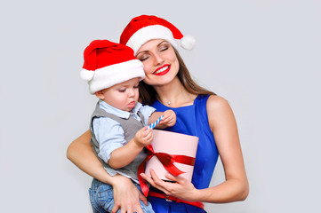 Mother holding her child and shows him a box with a red bow. A little boy takes out the gift from the box. Both in Santa hats