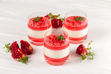 Creamy vanilla panna cotta with red jelly in beautiful glasses, fresh ripe pomegranate on white wooden background.