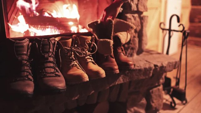 Woman Hand puts Winter Boots on a Shelf near Fireplace to get Warm. 4K. Vintage folk boots near the Cozy Fireside. Shoes getting dry near the burning fire at home, in chalet. Autumn and Winter Concept
