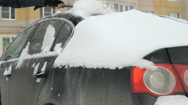 Caucasian man in casual clothes cleaning car from the snow near the house in winter against the backdrop of multi-storey residential building